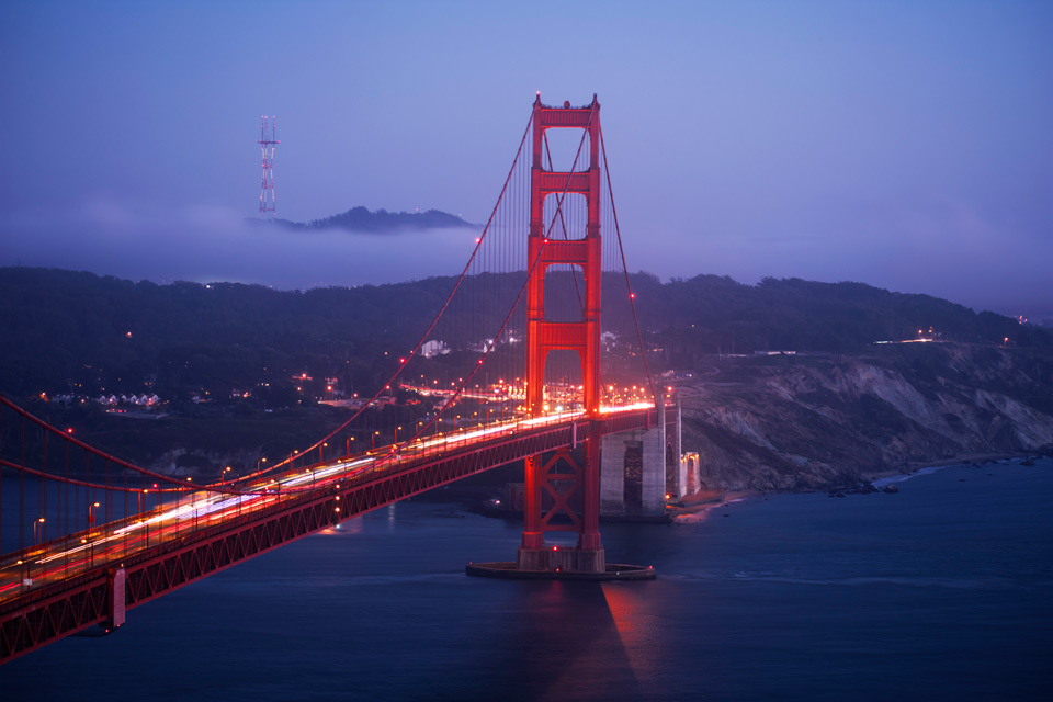 The Golden Gate Bridge, during the final moments of sunlight, with a great number of light trails along the surface of the bridge that are a product of car movement during a long exposure. A layer of fog and a tower also linger in the background.