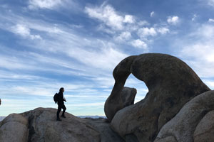 Young, free spiriting individual with a camera, standing in front of the famed Mobius Arch with a blue sky with thin cloud cover in the background.
